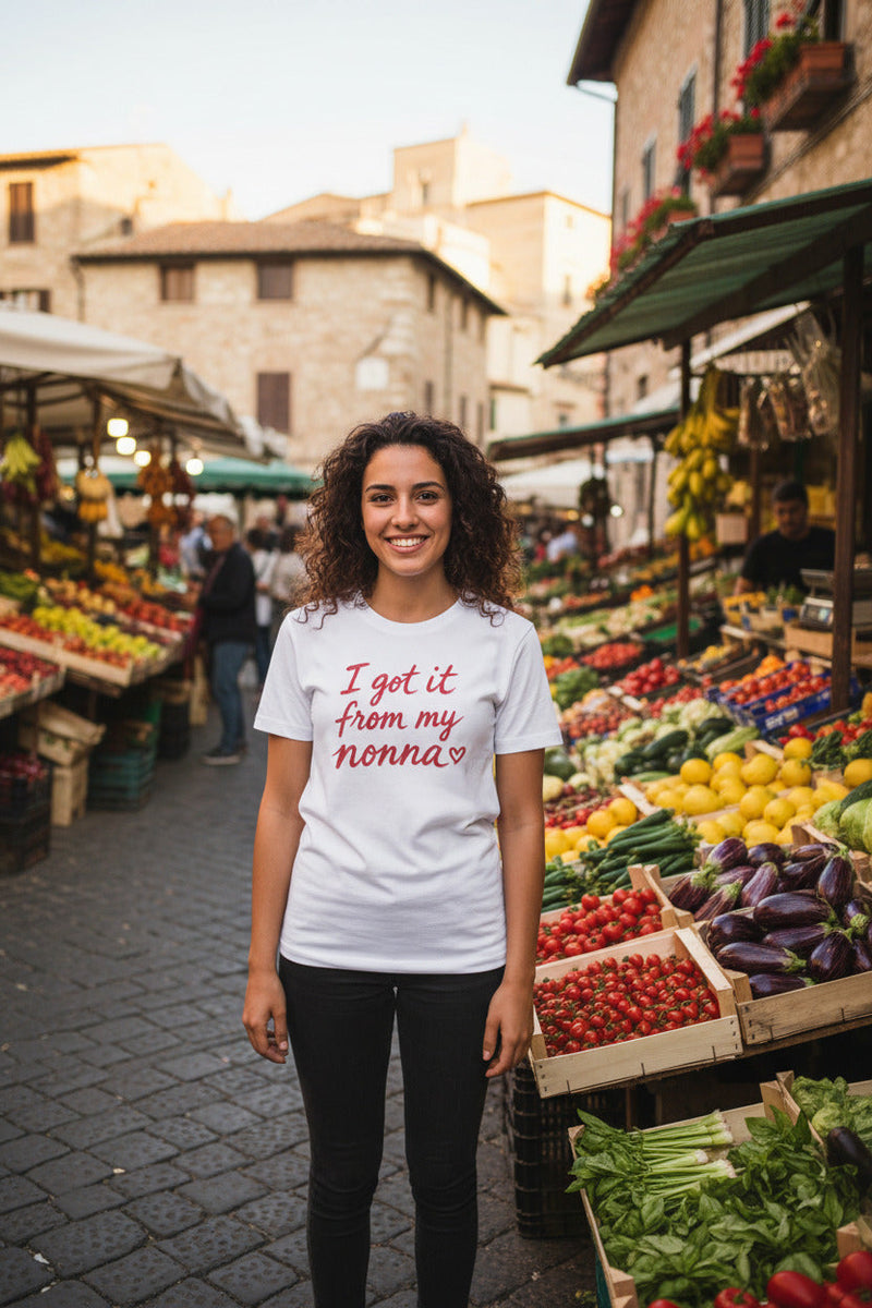 A white short-sleeve crewneck t-shirt featuring the phrase 'I got it from my nonna' printed in red cursive text across the chest, accompanied by a small red heart at the end.