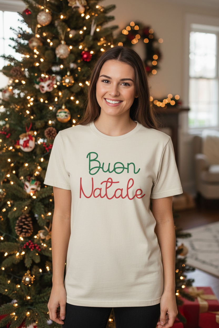 A woman wearing a cream-colored t-shirt with 'Buon Natale' printed on the front in green and red script, standing in front of a decorated Christmas tree in a cozy living room.