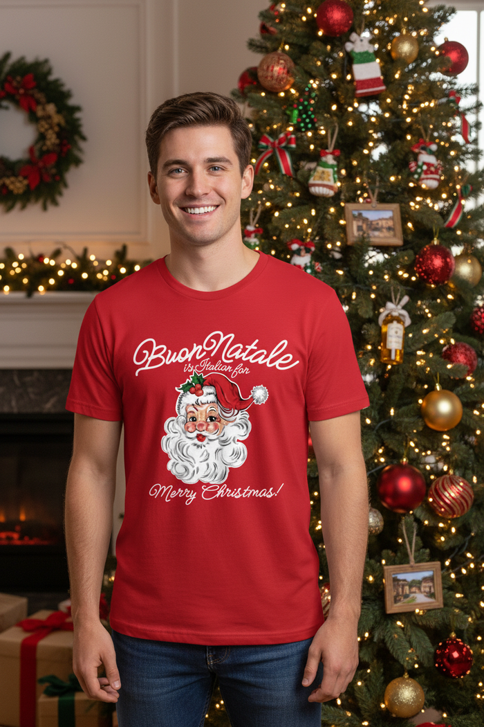 A smiling man wearing a red graphic t-shirt with 'Buon Natale' and a cartoon Santa Claus, standing in front of a decorated Christmas tree and fireplace.