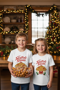Two kids in white Struffoli Squad shirts holding a bowl of struffoli in a Christmas-decorated Italian American kitchen