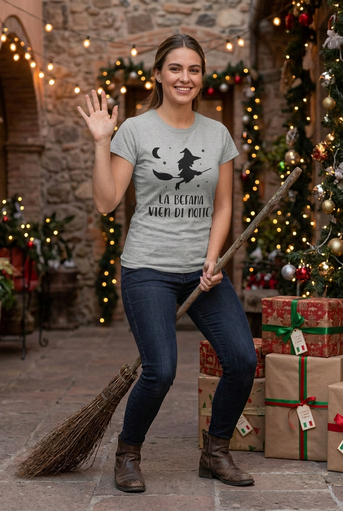 Woman in the gray Befana tee waving with her right hand while posing on a broom